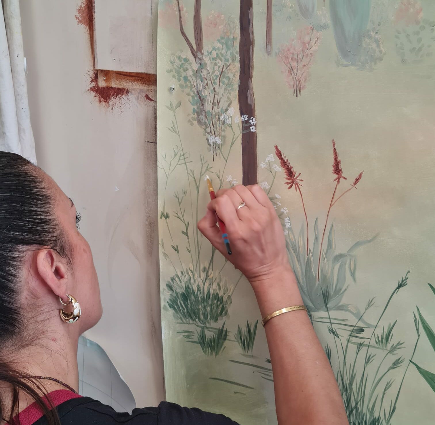 Artisane peintre décoratrice réalisant à la main une fresque murale végétale aux motifs de fleurs et de graminées.